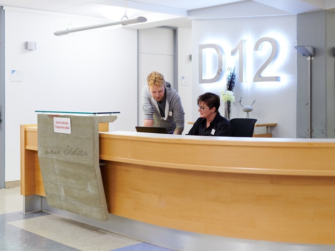 Anmeldung der Station D12 mit zwei Mitarbeiter:innen im Gespräch. (vergrößerte Bildansicht wird geöffnet)