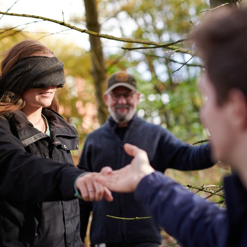 Ein junger Mann führt eine Frau mit verbundenen Augen an der Hand durch den Wald. Im Hintergrund ist Waldtherapeut Stefan Alberts zu sehen.