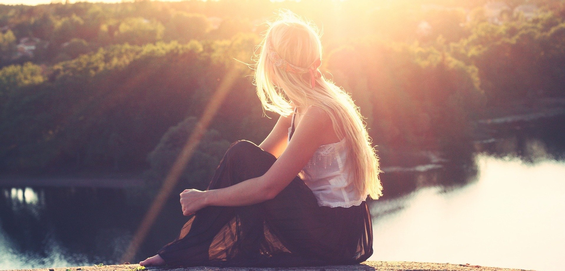 Eine junge Frau mit langen, hellblonden Haaren sitzt barfuß auf einem Felsen und schaut in die Sonne, die ihr Gericht direkt anstrahlt